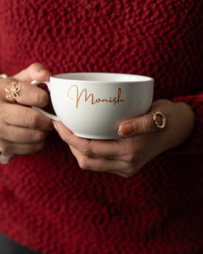 Weiße Keramiktasse mit Gravur „Monish“ in Goldschrift, von zwei Händen gehalten